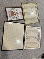 Four framed artworks viewed from above on carpet: two Basswood Centennial Tribute plaques, one engineering drawing, one fishing needlepoint canvas.