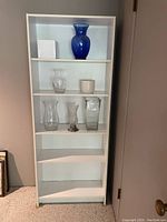 Full front photo of white tall bookcase with four shelves, some glass vases on shelves (not included).