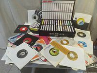 Large assortment of 45 RPM vinyl records scattered and stacked along with a black cassette tape case filled with tapes, some records in white paper sleeves.