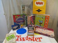 Photo showing seven board games including Rummikub, Cranium, Trivial Pursuit, Scrabble, a pack of playing cards in blue box, an official Scrabble dictionary, SAT flash cards, several party games books, and a partially visible Twister mat.