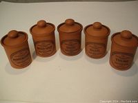 Five vintage terracotta pottery herb jars with tight-fitting lids and sealing gaskets, labeled peppercorn, mixed spice, sage, basil, and thyme, arranged in a row.