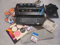 Full view of IBM Selectric II typewriter with surrounding accessories including typeface balls, ribbon cassettes, carbon paper, correction tapes, manual, dust cover, and base pad.