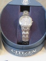 Ladies Citizen Eco-Drive watch displayed in original Citizen round box with navy blue interior and lid, showing stainless steel bracelet and silver-tone case.