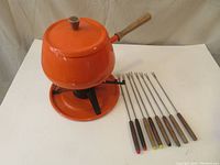 Full shot of orange fondue pot with wooden handle and lid on its black metal stand and circular base plate, alongside eight fondue forks with wooden handles, showing two forks missing colored tips.