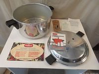 Photo showing the main aluminum pressure canner pot, lid with black handles, perforated steaming rack, and instruction manuals laid out on a table.