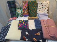 Photo showing 16 assorted fabric pieces in folded stacks featuring various prints including plaid, floral, travel-themed, and bird prints, plus pink plush fabric.
