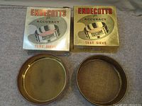 Two vintage Endecotts test sieves and their original boxes placed on carpeted floor showing one sieve with fine mesh and one with coarse mesh.