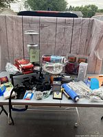 Multiple camping and utility items displayed on a table against a wood panel backdrop including lantern, trailer lights, and hardware parts.