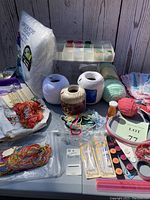 Craft items including various yarns, embroidery threads, needles, buttons, and some fabric and sewing accessories arranged on a table against a wooden panel background.