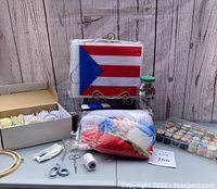 Full lot overview showing lace spools, Puerto Rican flag needlepoint project, container of vintage threads, two pairs of scissors, white pins, and other crafting supplies on a gray table with rustic wooden backdrop.
