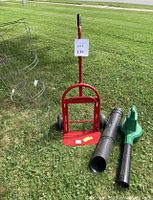 Red metal dolly with wheels and handle, two-piece green and black leaf blower attachments, and three wire garden tomato cages standing on grass.