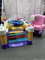 Front view of colorful Fisher-Price piano walker with blue sliding seat, keys visible, and pink Laugh & Learn chair behind