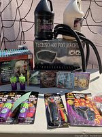 Photo showing Techno 400 fog machine box, two fog liquid jugs, red LED mist vampire skull box, purple LED pumpkin lights, pumpkin carving kits, power poker set, and Halloween-themed CDs all arranged on a table.
