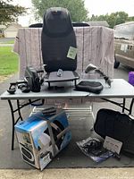Wide view showing all items on and under table including chair massager seat, foot/calf massager, handheld massager, portable minibike pedal exerciser in box, and vibration plate.