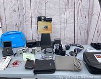 Wide view of various cameras, electronic devices, cables, cases, and accessories arranged on a gray folding table against a wood plank backdrop