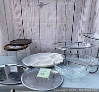 View of entire lot including wooden cheese board with tools to left, white embossed oval serving dish center right, aluminum platters, and plastic buffet server trays stacked on a metal frame stand in background.