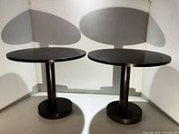 Front view of two round dark wood end tables with cylindrical supports and round bases, showing the tops and bases clearly.