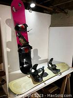Photo showing the two snowboards on a wooden table and against a white wall. One snowboard is upright with pink and black design, the other is lying flat with beige design.