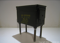 Front view of the ammo can mini grill showing markings on the side and four legs that are bolts or rods attached to the base.