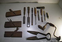View of the full lot showing three wooden hand planes, eight chisels and a hand drill brace on a white surface.