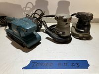Three power sanders on a table: one blue electric belt sander and two round orbital sanders, photographed with a note 'TESTED 8 15 23'