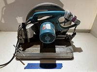 Makita cut off saw front and side views on metal base, showing overall tool condition and taped broken safety thumb switch on handle.