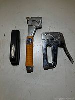 Three staplers on a white surface: one black plastic desktop stapler, one silver metal heavy-duty stapler with orange grip, and one silver metal staple gun with lever handle.