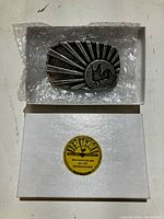 Photo of metal Sun Studios belt buckle on bubble wrap, above the white box featuring a yellow Sun Records logo and web address.