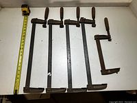 Five vintage metal clamps with wooden handles arranged in a row on a white surface next to a tape measure for scale.