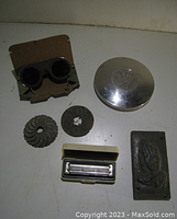 Photo of vintage miscellaneous items including welding glasses, VW hubcap, cast iron decor pieces, harmonica, and prayer hands plaque.