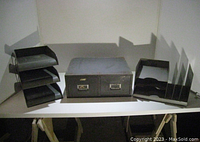 Photo of three vintage metal office desk items on white table against white background, showing a 3-tier letter tray, 2-drawer filing box, and 4-section file sorter rack.