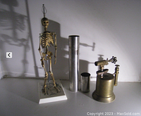 Vintage anatomical skeleton, brass blow torch, and two metal cylindrical containers arranged together on a surface.