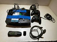 Photo showing the original Xbox console with blue tape holding down the disc tray, two wired controllers, a remote control, AV cable, and a USB dongle all laid out on a surface.