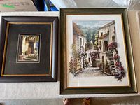 Two framed art prints on table, one smaller with dark frame and smaller artwork, other larger with greenish frame and larger artwork showing stucco houses and flowers.