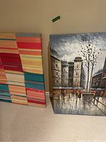 Two large canvas paintings leaning against a wall: one colorful abstract stripes, one city street scene