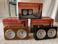 Five analog chess clocks arranged with their original orange chess-themed boxes behind them. Two clocks visible in front, three boxes visible behind.