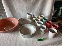 Photo shows large Pyrex mixing bowl, 6 floral rim plates, 2 sets of bowls, and 10 assorted mugs in varied colors and prints arranged on table.