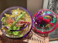Two Perplexus 3D maze puzzle balls shown resting on a chair, one larger with yellow and green internal maze labeled 'Original' and a smaller colorful one.