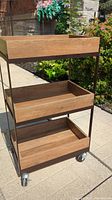 Front view of 3-tier utility cart showing chestnut finish wooden shelves and brown metal frame on caster wheels outside on concrete.