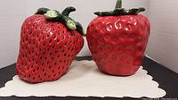 Two ceramic cookie jars shaped like large red berries with green leafy lids, displayed on a white cloth.