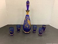 Full view of the cobalt blue decanter with stopper and six matching small glasses arranged in front of a white backdrop.