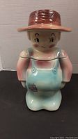 Front view of the ceramic cookie jar shaped like a boy wearing a brown hat and blue overalls with a patch.