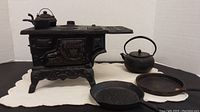 Miniature black cast iron stove with pots, kettle, and frying pans arranged on white cloth.