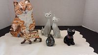 Six cat figurines arranged on a white surface: a large orange/white ceramic cat, two white ceramic cats, a small black ceramic cat with a red collar, a mosaic styled metal cat, and a small shiny metal rounded cat figure.