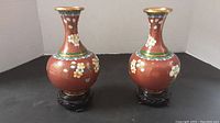 Two red cloisonné vases on wooden stands, showing front views with floral designs and gold rims.