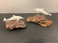 Front angle view of two John Perry dolphin decor pieces showing white dolphins on irregular natural wood bases with plaques.