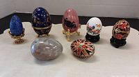 Photo of seven decorative eggs of varying materials and colors arranged in a row, showing details and stands.