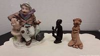 View of three figurines from the front on a white cloth surface against a white background: porcelain man on bench, black wood man kneeling, and clay couple standing.