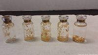 Five small glass vials containing 24K gold flakes arranged in a row, showing varying amounts of flakes inside and labeled with gold bands around the necks.