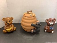 Honey pot cookie jar with attached black bear climbing and two ceramic bear cookie pots with lids arranged side by side